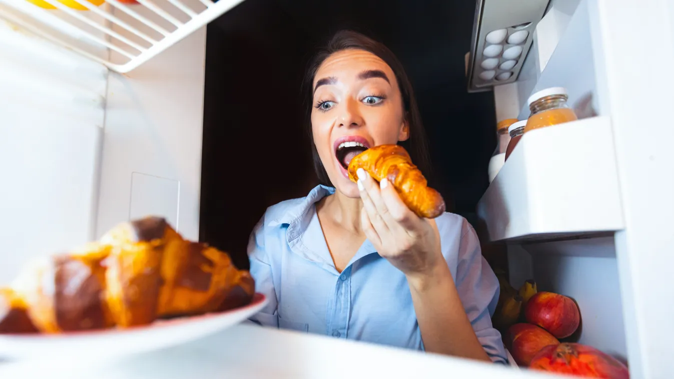Csalónapot tartó nő eszik egy croissantot a hűtőből.