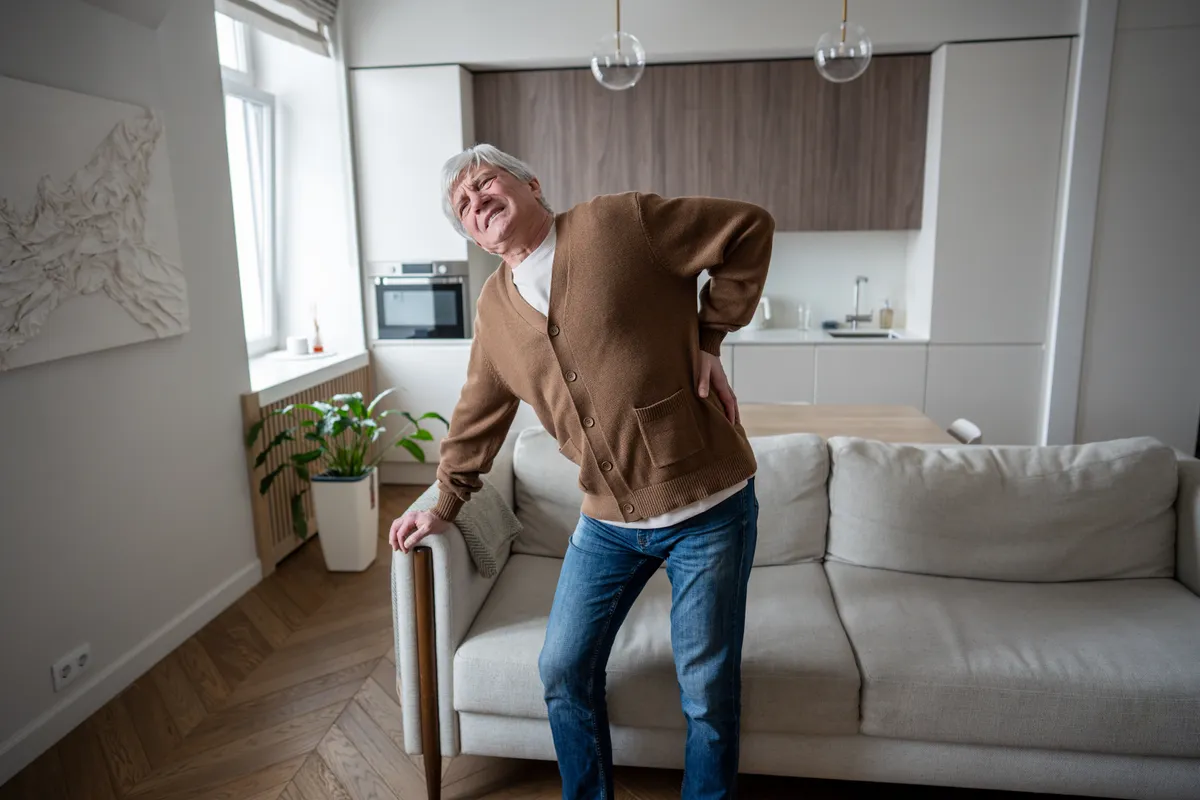 Csigolyaelcsúszással küzdő idős férfi a hátát fájlalva a nappalijában