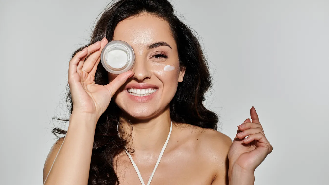 Arckrémet a jobb szeme elé tartó boldog fiatal nő tavaszi bőrmegújítás közben