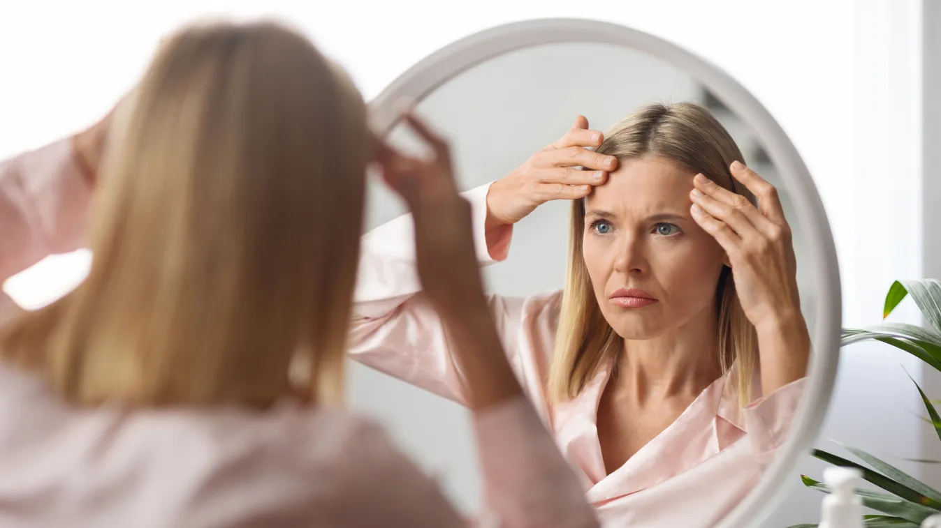 Tükörben a homlokát vizsgáló nő ránctalanításon gondolkodva