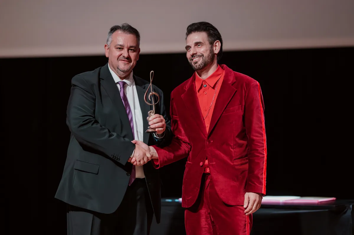 Rácz Gergő kezet ráz a díjátadón, miközben átveszi az elismerésnek járó trófeát.