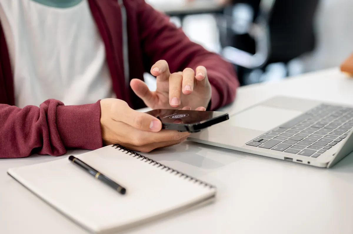 Mesterséges intelligencia az oktatásban: egy diák kezében mobiltelefon, előtte laptop, jegyzetfüzet és toll.