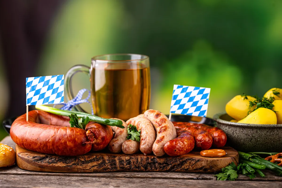 Bajor kolbászok, zöldségek, burgonya, pohár sör egy fatálon, mellette kicsi bajor zászlók.