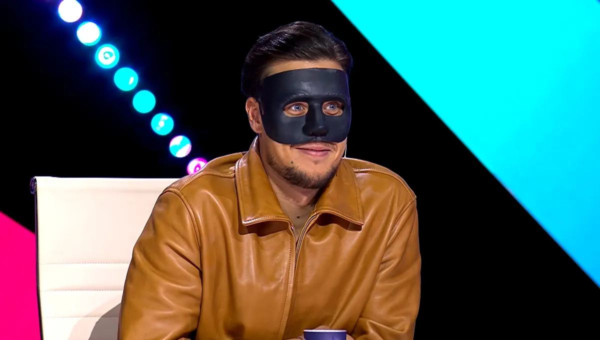 T. Danny fekete maszkban és világosbarna bőrdzsekiben néz megilletődve a zsűriasztal mögül.
