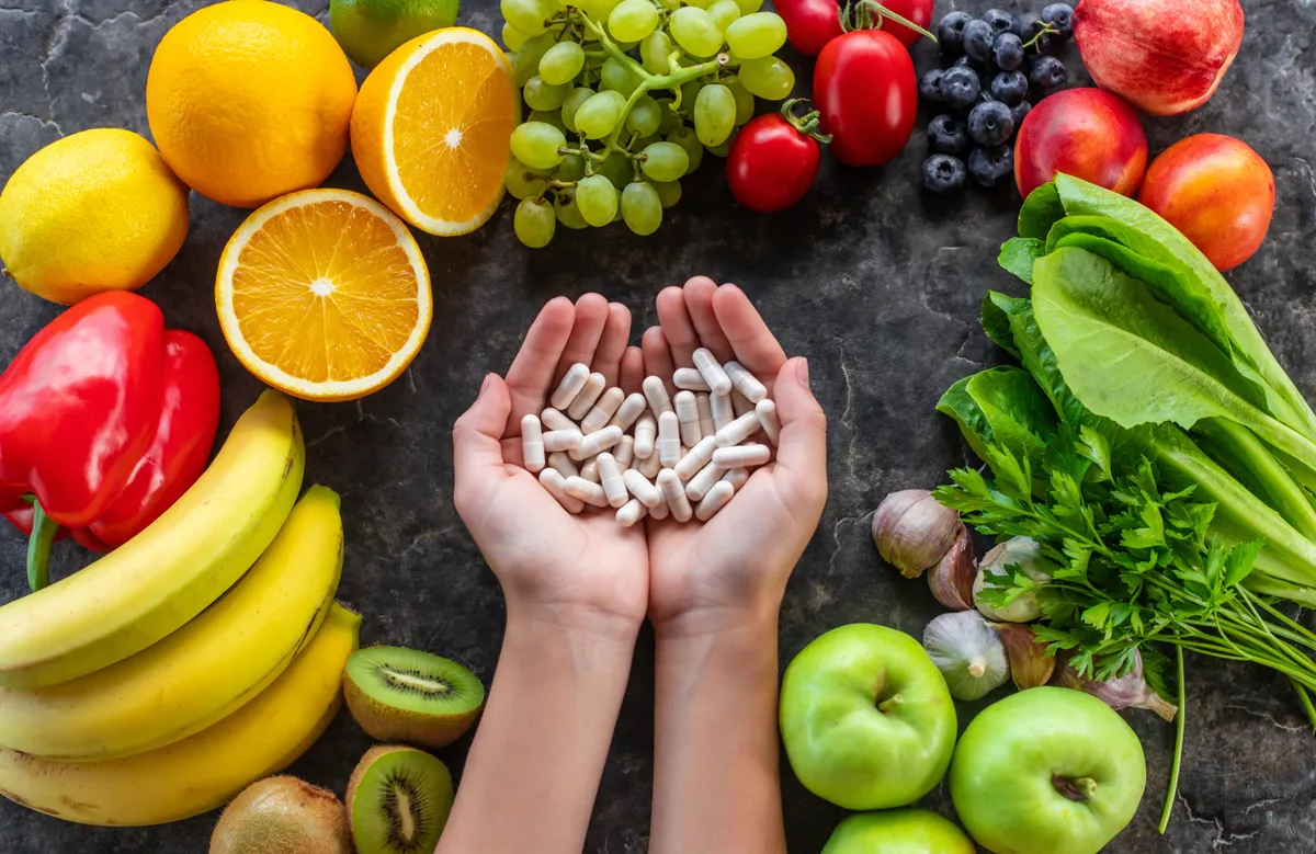 C-vitamin tartalmú zöldségek és gyümölcsök, középen egy nő tenyerében vitamin kapszulák